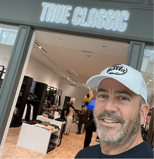 Man in a white cap smiling outside a store named 'True Classic.'